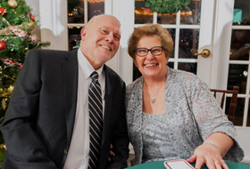 Marjorie Bryner and another person sit at a table during a holiday event. A white door and Christmas decorations are behind them.